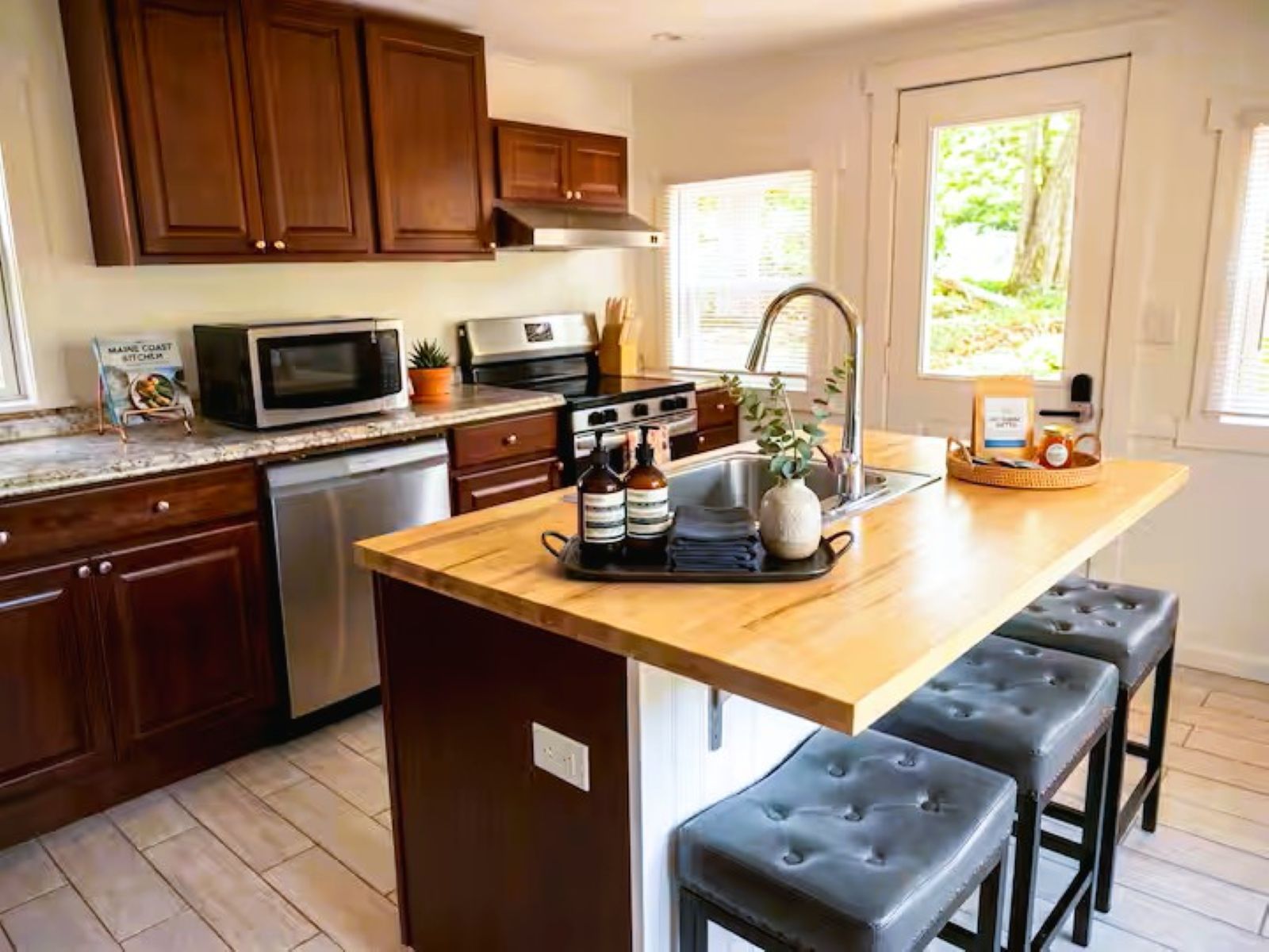 A kitchen with wooden cabinets, a microwave, a stove, and a sink. There are two stools and a tray with bottles and a plant on the island.