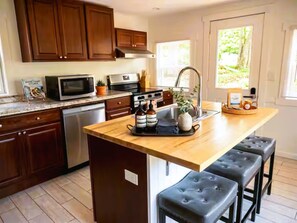 A kitchen with wooden cabinets, a microwave, a stove, and a sink. There are two stools and a tray with bottles and a plant on the island. - Spacious Peaceful Redding Cottage Retreat (Redding)