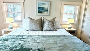 Ein Schlafzimmer mit einem Bett, zwei Nachttischen, einem gerahmten Bild an der Wand und zwei Fenstern.