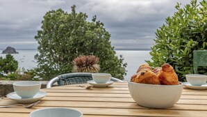 Ein Tisch mit einer Schale Gebäck, zwei Tassen und ein Stuhl mit Blick auf das Meer und Bäume.