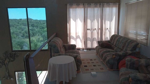 A living room with a sofa set, a small table, and a view of a forest through large windows. - Private farmhouse in the bush with AC, just outside Windhoek on a game farm. (Windhoek)