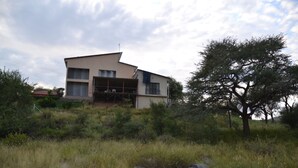 A modern two-story house with a sloped roof, surrounded by natural vegetation and a large tree. - Private farmhouse in the bush with AC, just outside Windhoek on a game farm. (Windhoek)