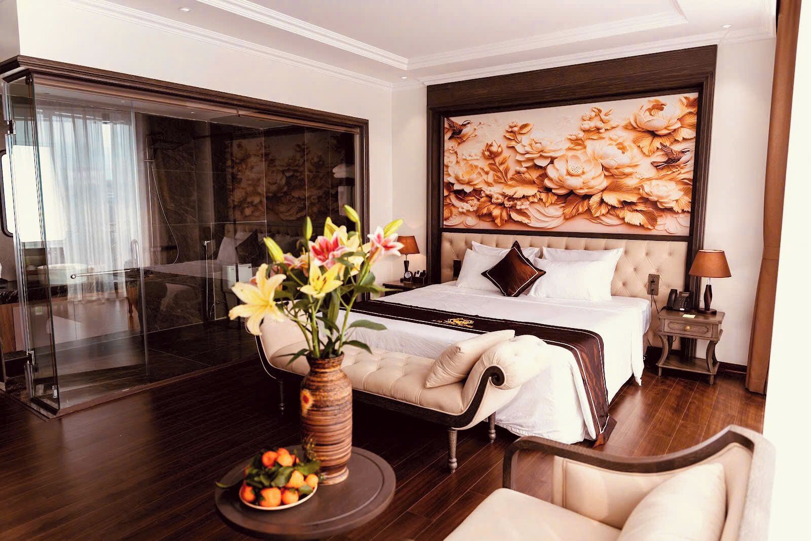A hotel room with a bed, a bedside table with a lamp, a vase with flowers, a chair, and a large window with curtains.