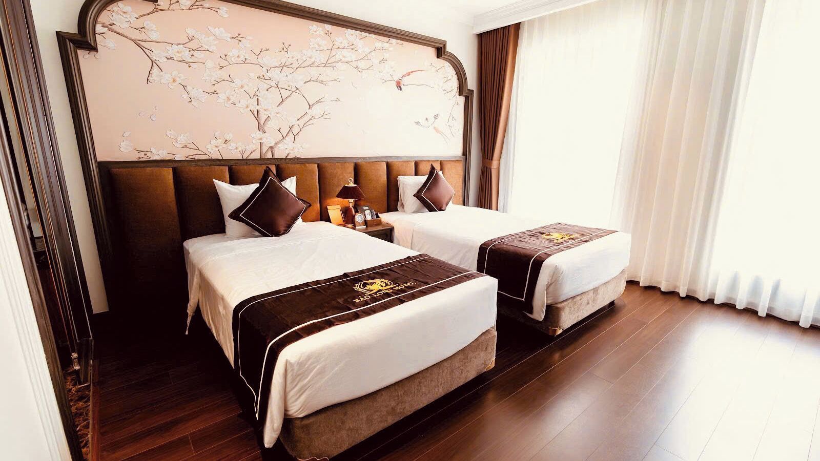 A hotel room with two beds, a large wall mural, a wooden floor, and a window with curtains.