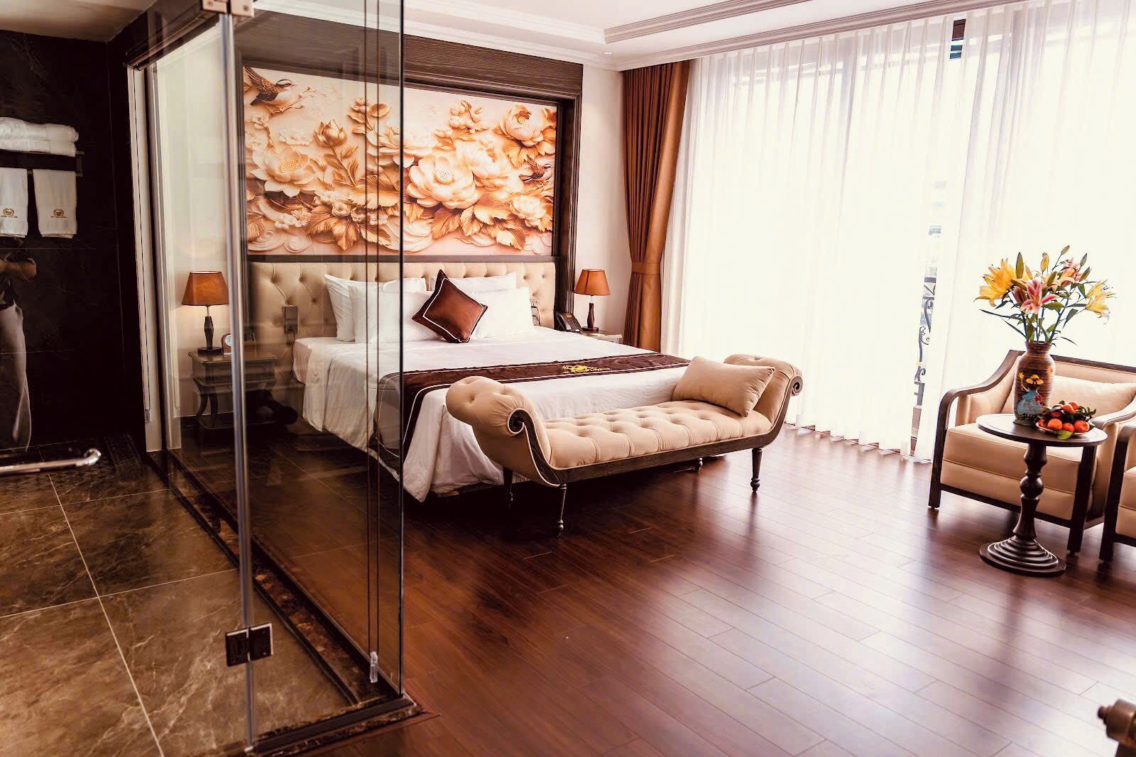 Una habitación de hotel con una cama grande, un separador de vidrio, un sofá, una pequeña mesa con un jarrón y una silla.
