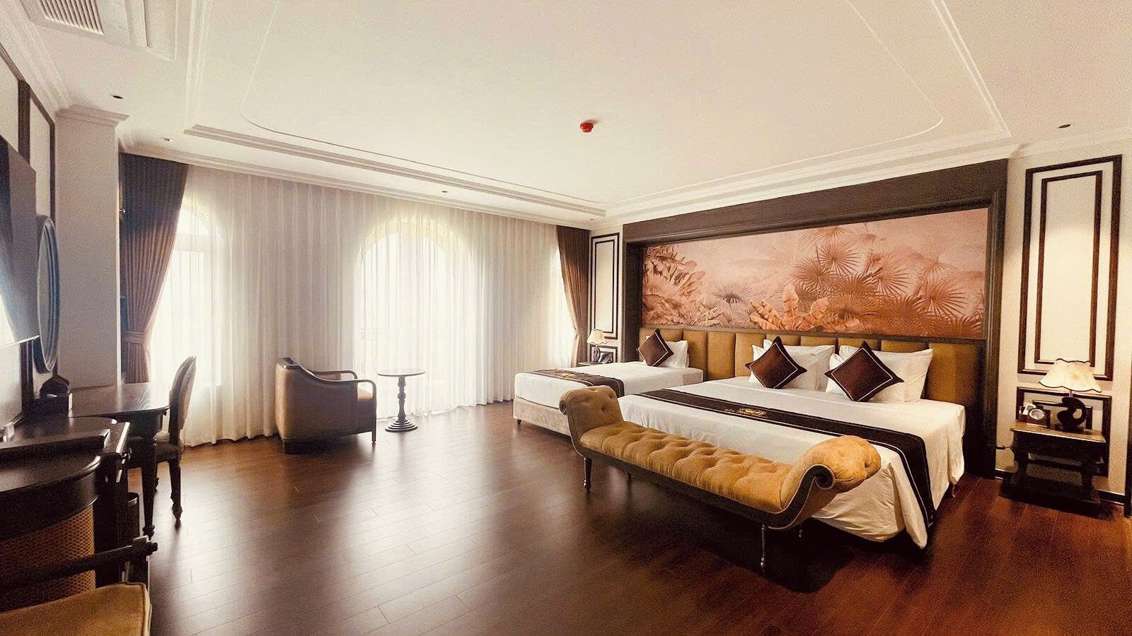 A hotel room with a large bed, a sitting area with a bench, a desk, and a large window with curtains.