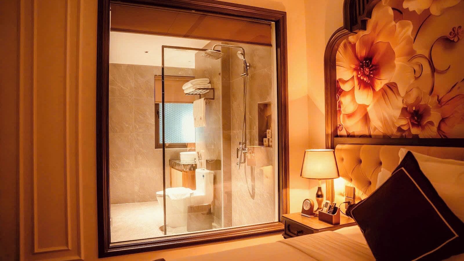 Habitación de hotel con ducha de vidrio, cama con cabecera, mesita de noche con lámpara y decoración floral en la pared.