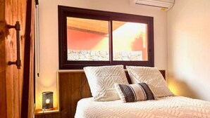 Ein Schlafzimmer mit Bett, zwei Kissen, einem Nachttisch, einer Lampe und einem Fenster mit Blick auf einen Sonnenuntergang.