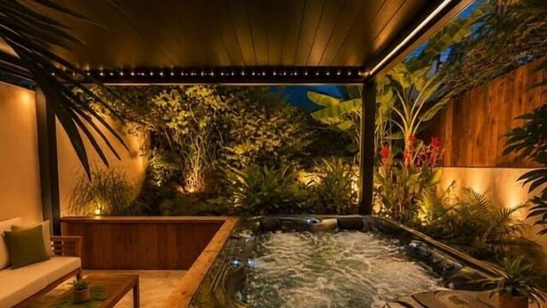 A hot tub in a covered outdoor area with wooden flooring, surrounded by lush greenery and a wooden wall. - Tiny house with private Jacuzzi (Saint-Paul)