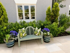 A green bench with patterned cushions, two potted plants with blue flowers, and a window with white frames. - The Station at Shustoke (Birmingham)