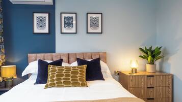A neatly made bed with pillows, a bedside lamp, a nightstand with a plant, and framed artwork on the wall.