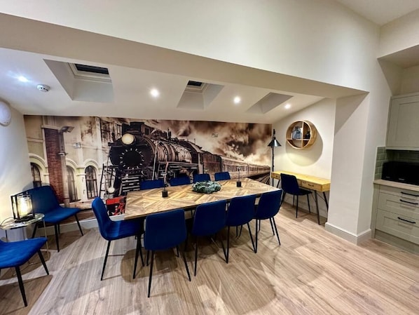 A dining area with a large table, blue chairs, a mural of a train, and a modern kitchen in the background. - The Station at Shustoke (Birmingham)