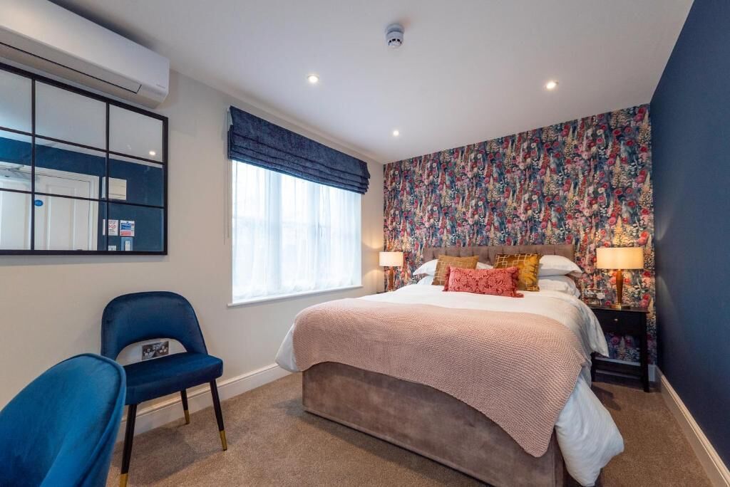 Un dormitorio con una cama, una silla, una mesita de noche, un ventanal con cortinas y una pared con estampado floral.