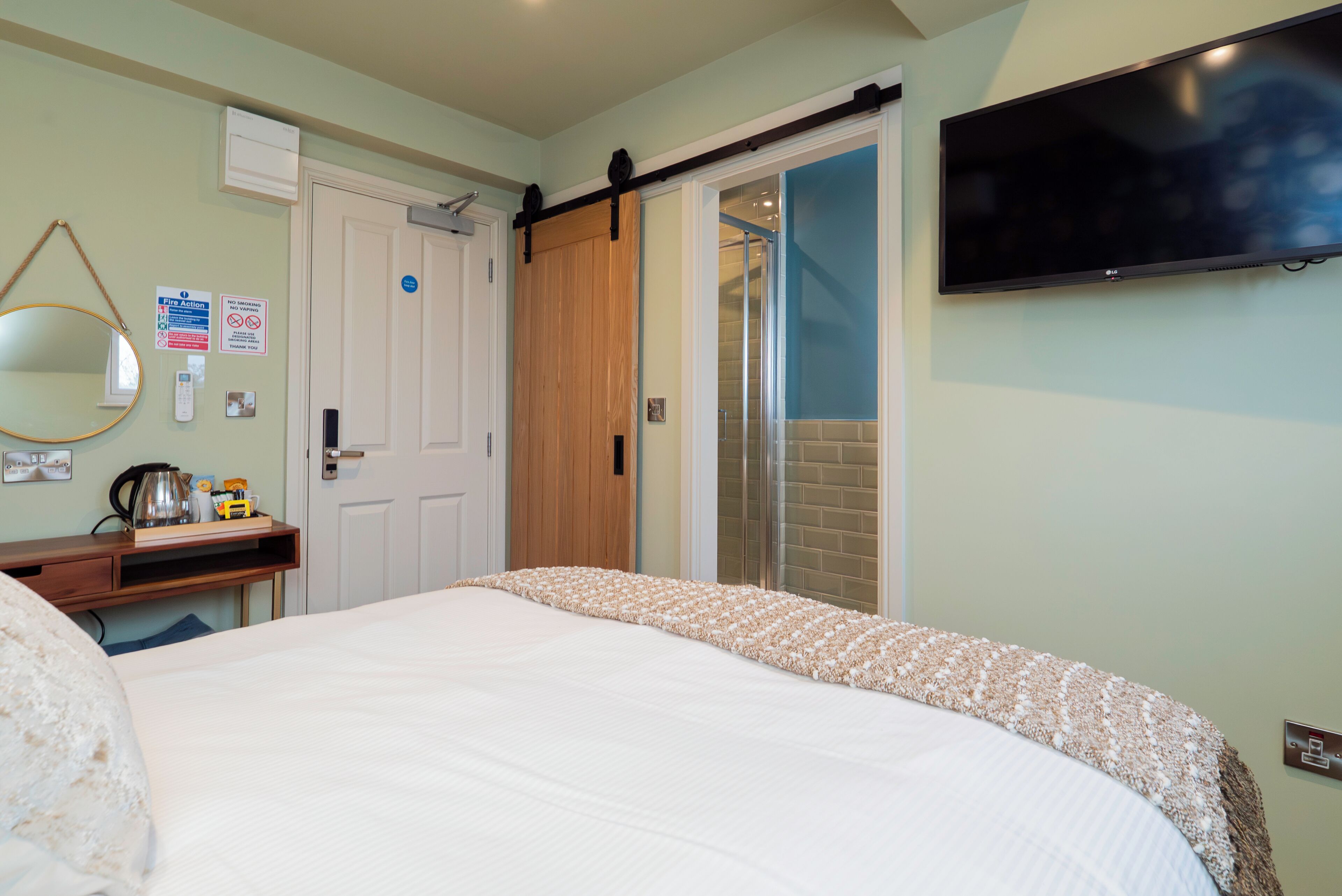A bedroom with a bed, a desk, a mirror, a TV, and a sliding door leading to a shower area.