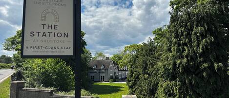 A sign for The Station, a first-class stay at Shustoke, with a view of a traditional house and lush greenery.