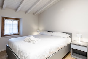 Quarto com cama, mesas de cabeceira, janela com cortinas e teto com vigas de madeira.
