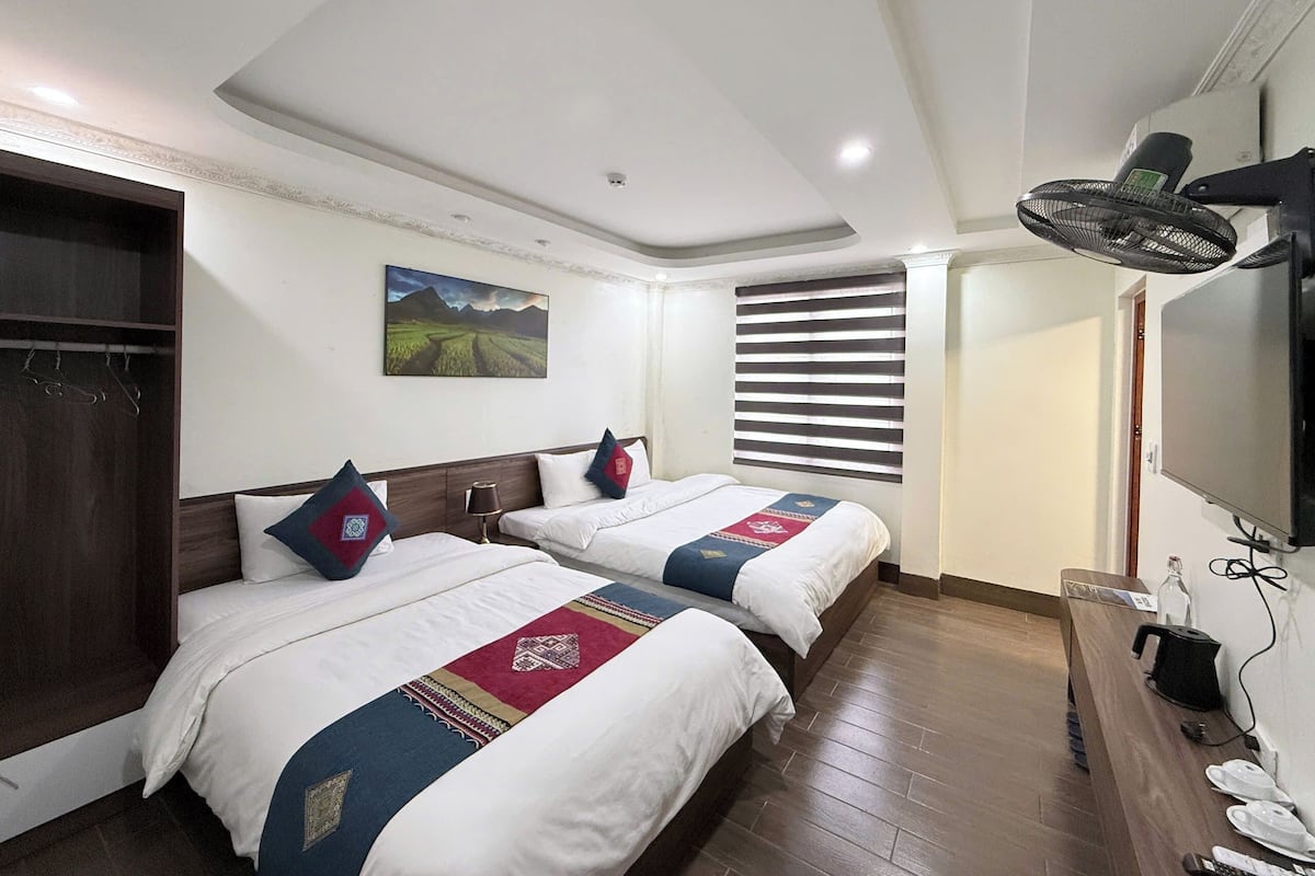 Habitación de hotel con dos camas, un armario de madera, un cuadro en la pared, un ventilador de techo y un televisor montado en la pared.
