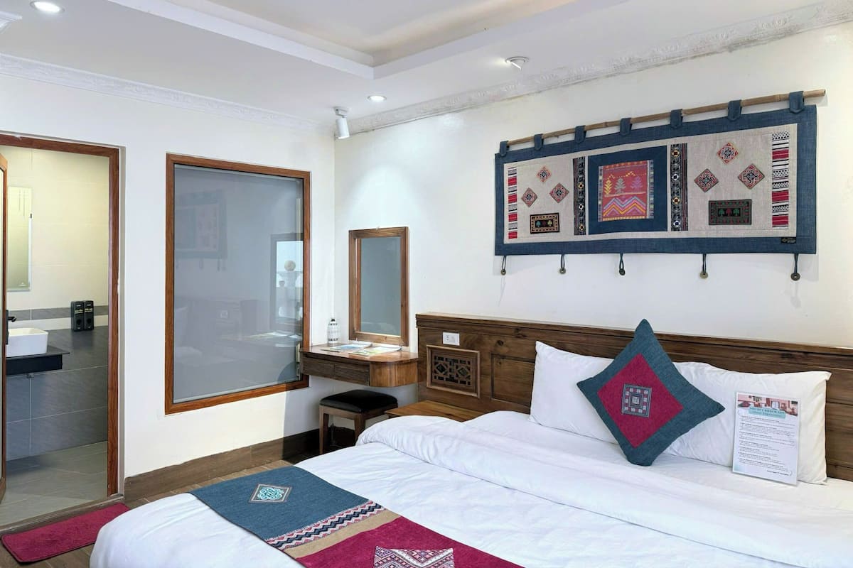 Habitación de hotel con una cama, una mesita de noche, un espejo, un tapiz colgado en la pared y una puerta que da al baño.