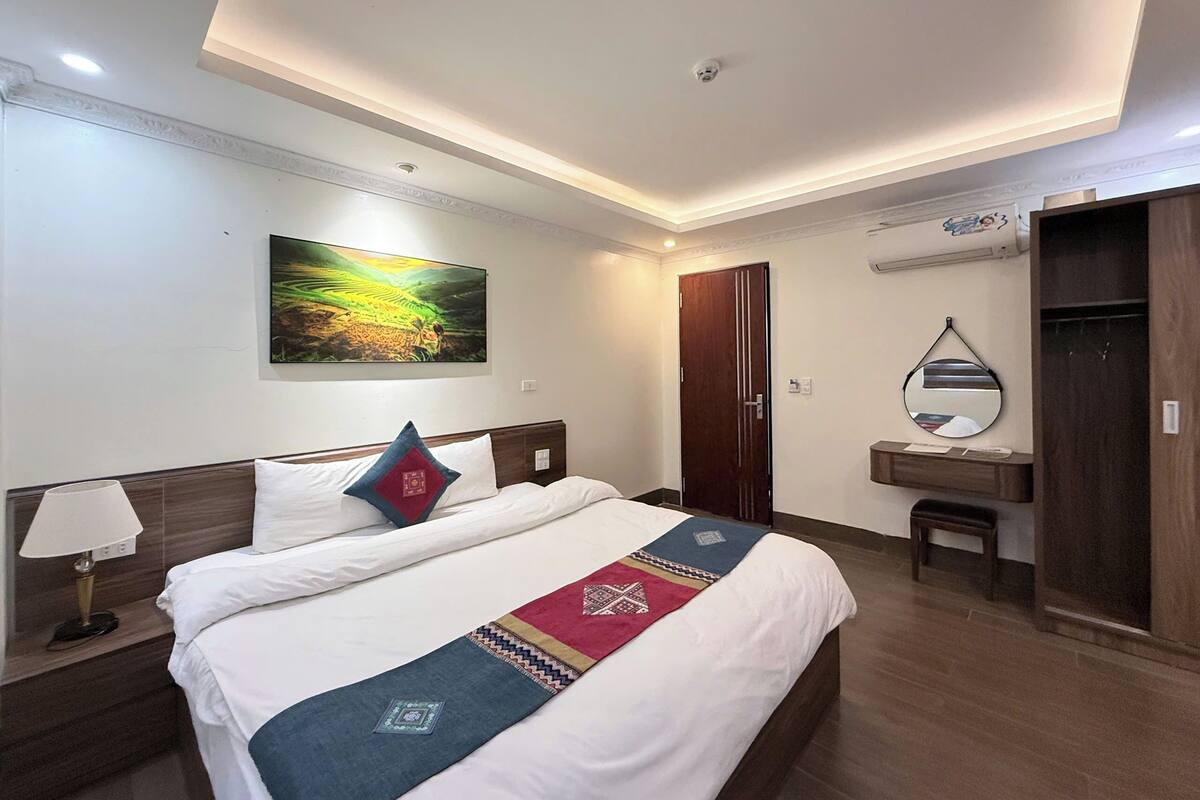Habitación de hotel con cama, mesita de noche, escritorio, espejo y un cuadro en la pared.