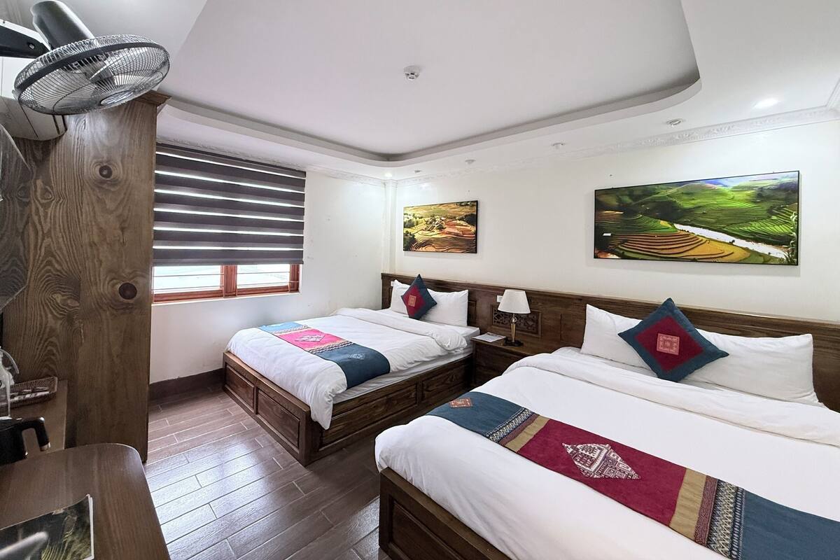 Habitación de hotel con dos camas, piso de madera, ventilador de techo y dos cuadros de paisajes.