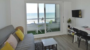 A room with a sofa, a small table, a TV, a balcony with a view of the sea, and a chair. - Window overlooking the sea, loggia overlooking the sea on the beachfront (Courseulles-sur-Mer)