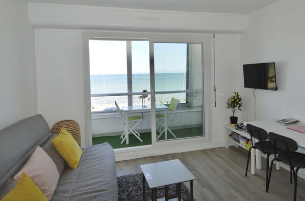 Quarto com sofá, mesa pequena, televisão, varanda com vista para o mar e uma cadeira.