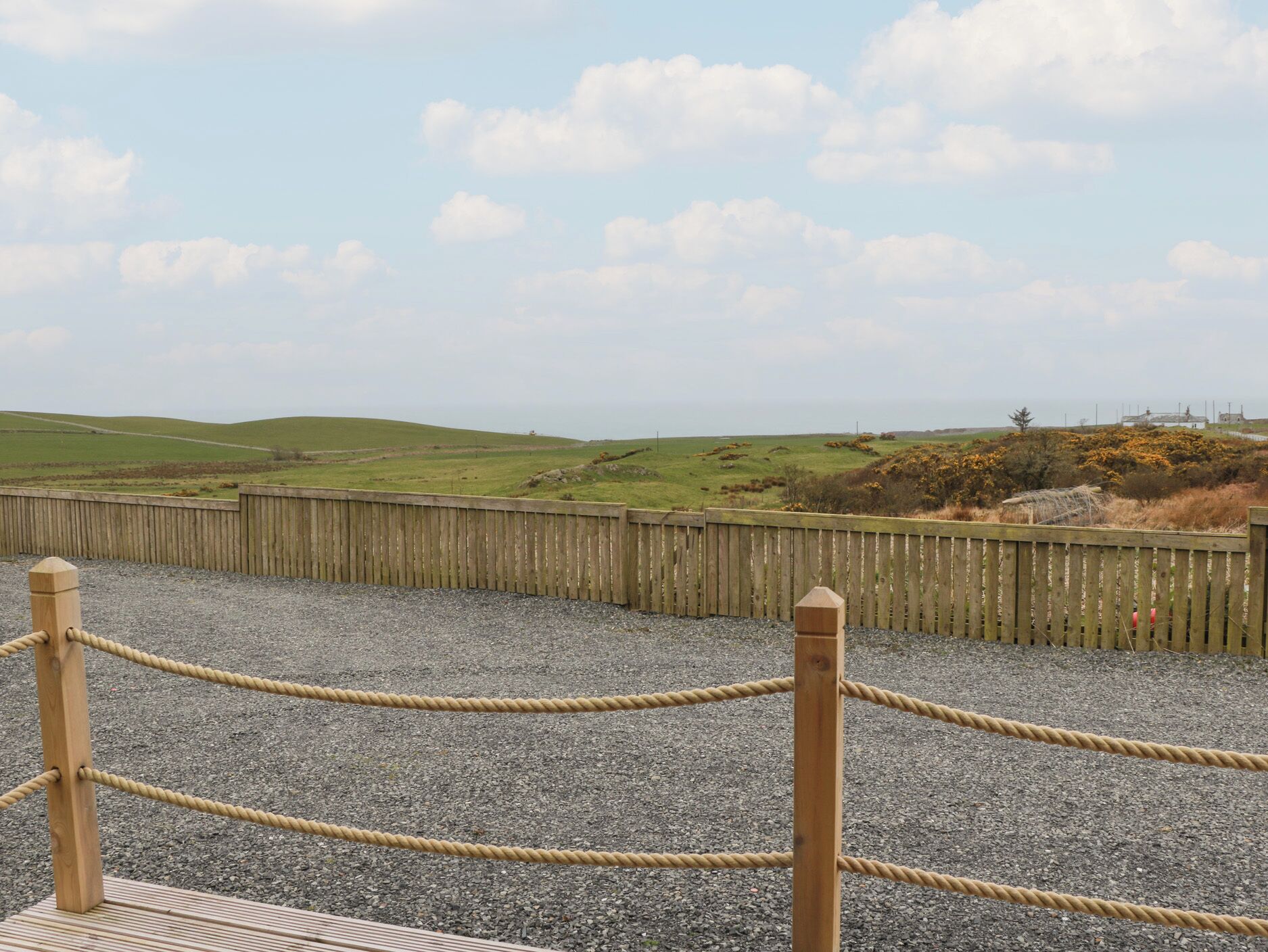 A wooden fence with a rope barrier, gravel ground, and a view of green hills and a cloudy sky.
