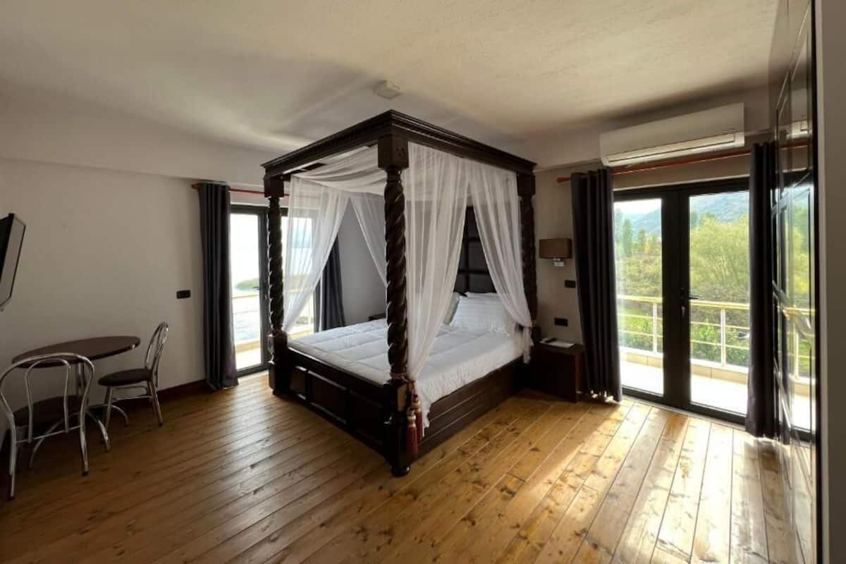 Ein Bett mit Baldachin, ausgestattet mit weißer Bettwäsche, Holzboden, ein kleiner Tisch mit Stühlen und Blick ins Grüne durch große Fenster.