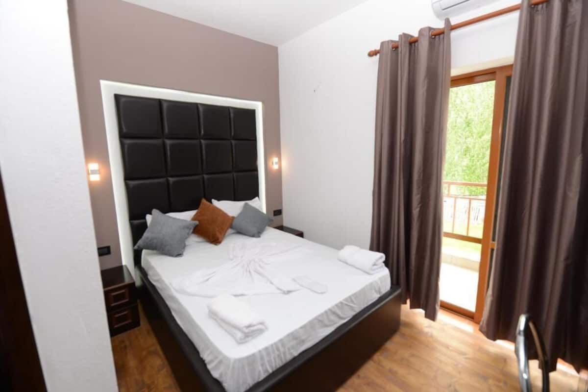 Ein Schlafzimmer mit Bett, Kopfstütze, einem Fenster mit Vorhängen und Blick auf Grünfläche.