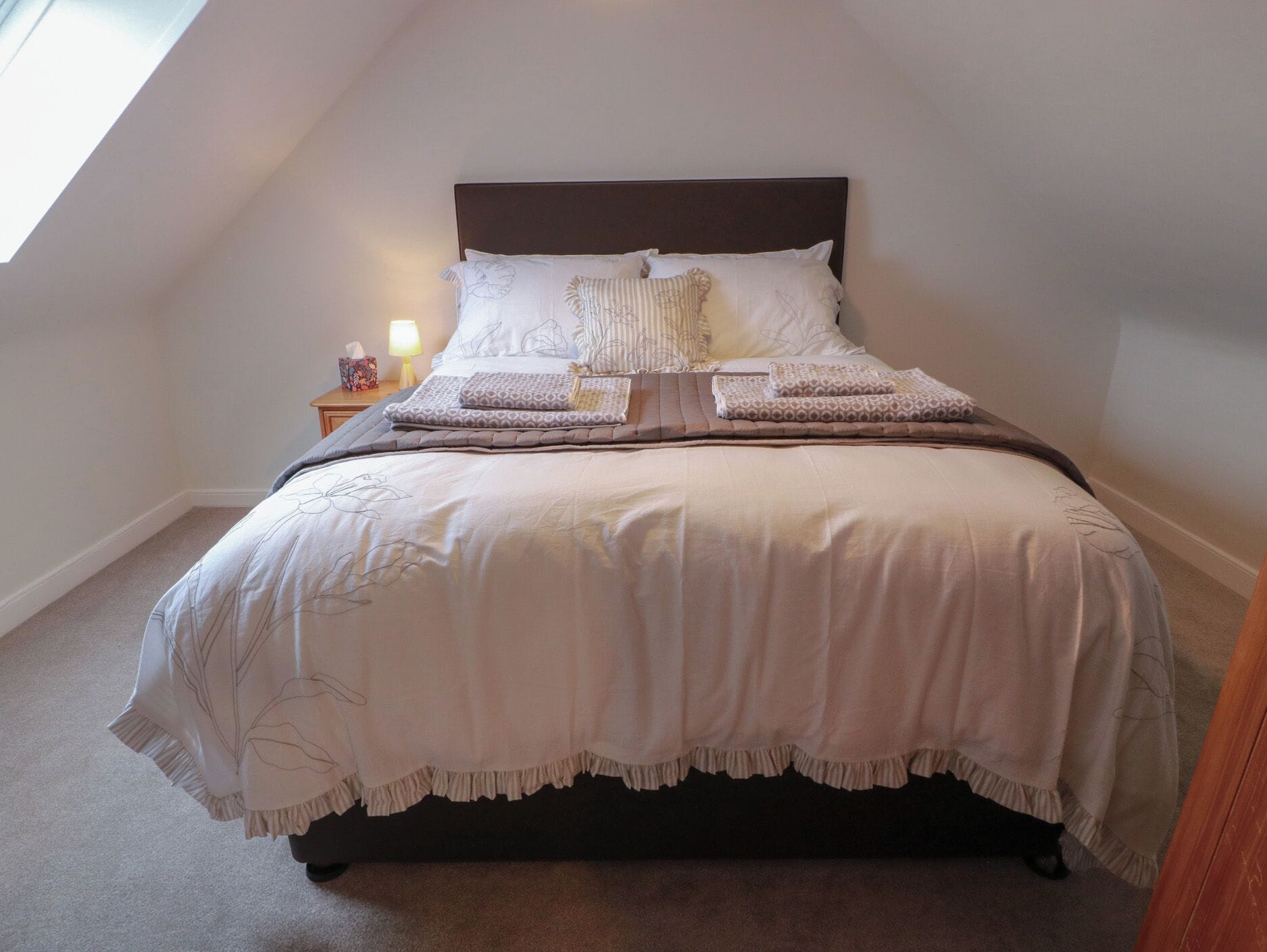Ein ordentlich bezogenes Bett mit Kopfstütze, Kissen und Decke in einem Raum mit geneigter Decke und einem Nachttisch mit Lampe.