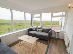 A room with a grey sofa, a small table, and a view of a green landscape through large windows. - South Kirklauchline Farm (Stranraer)