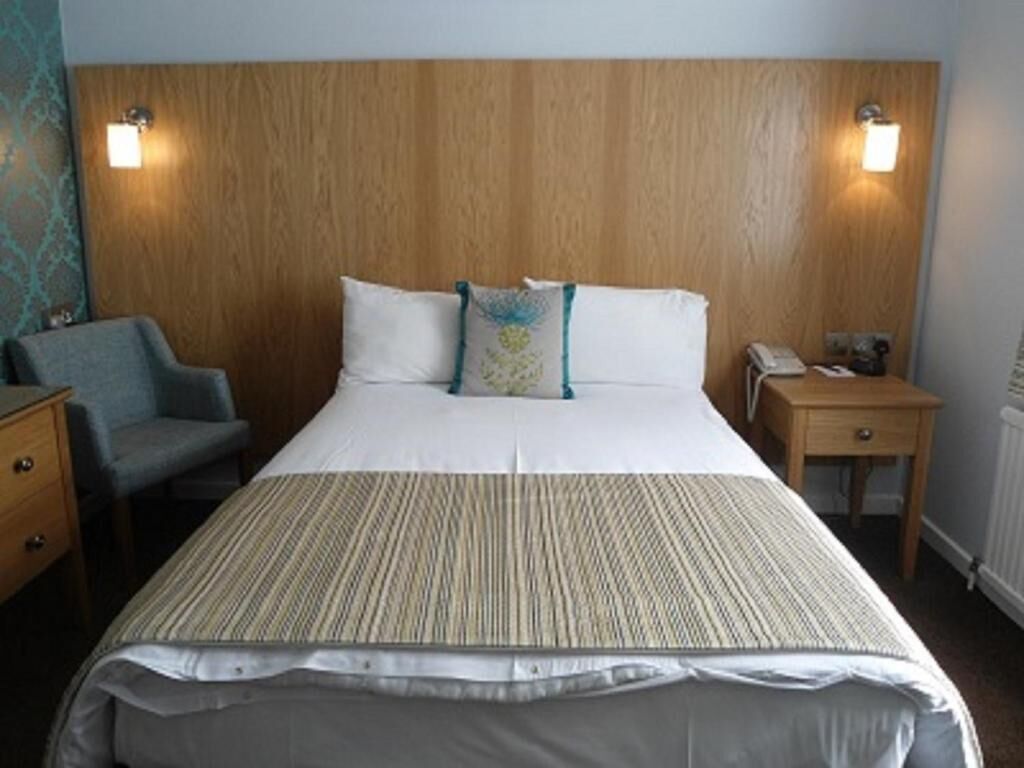 A hotel room with a wooden headboard, a bed with white linens, two bedside lamps, a chair, a nightstand, and a small table.