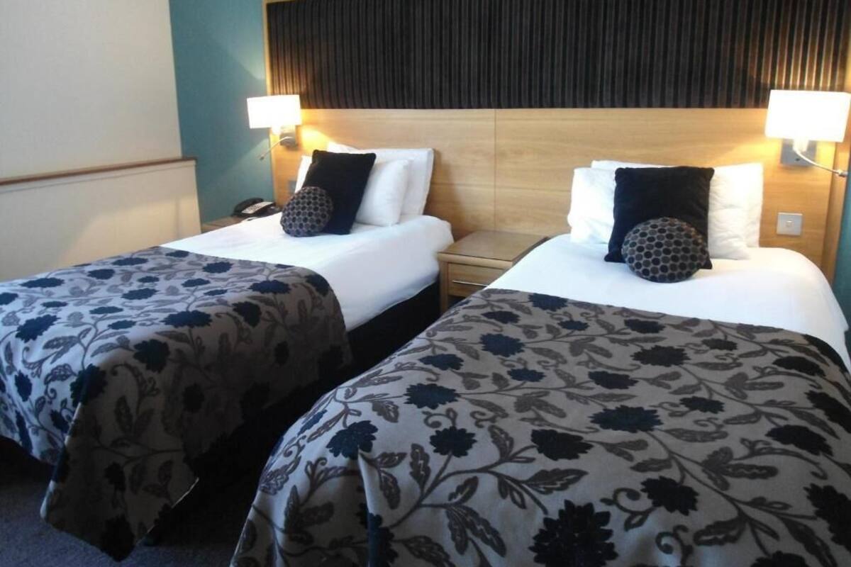 Una camera d'albergo con due letti, ognuno con una coperta a motivi floreali, un poggiatesta in legno e lampade da comodino.