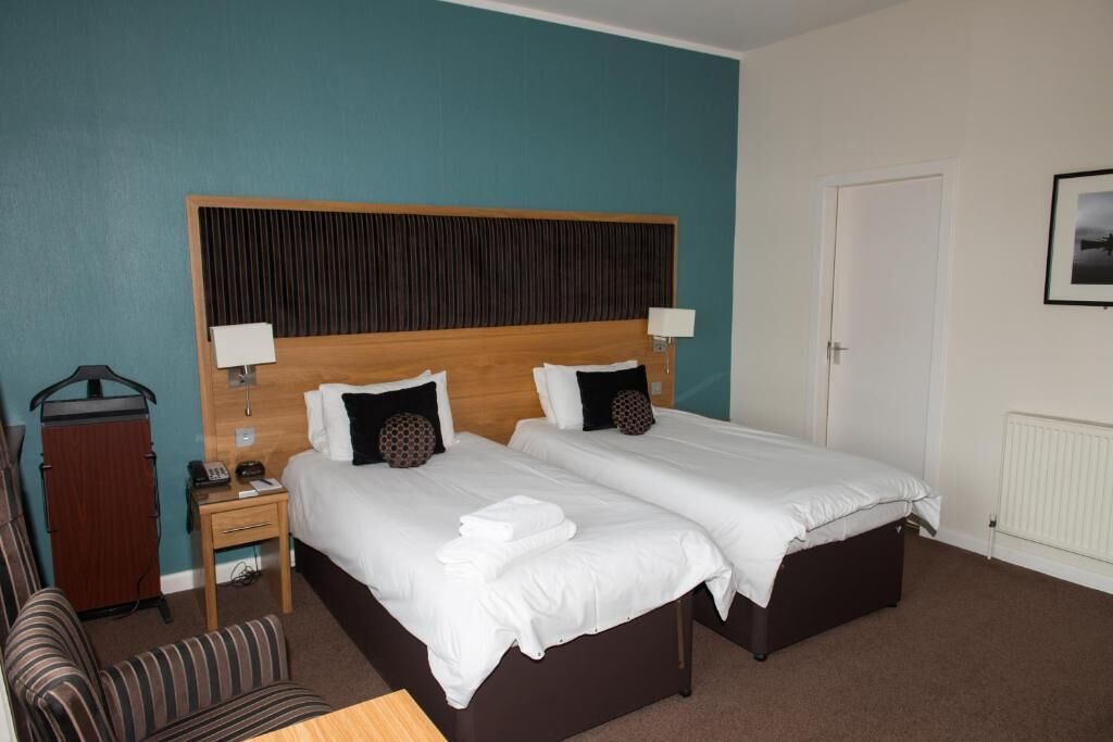 Habitación de hotel con dos camas, cabecero de madera, mesita de noche con lámpara, una silla y perchero.