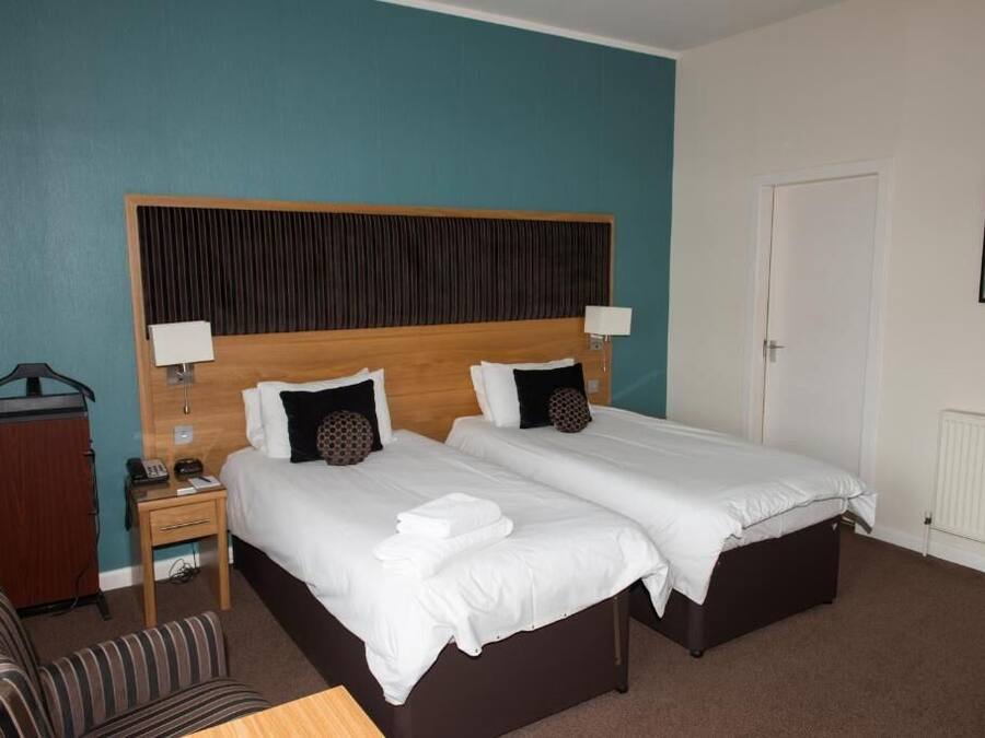 Habitación de hotel con dos camas, cabecero de madera, mesita de noche con lámpara, una silla y perchero.