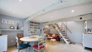 A dining area with a round table and chairs, a staircase with a railing, and a living area with a sofa and cushions. - Gîte "Gite De La Rampotiere" with Private Terrace, Garden, and Wi-Fi (Quettreville-sur-Sienne)