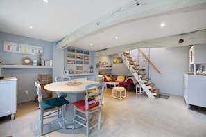 A dining area with a round table and chairs, a staircase with a railing, and a living area with a sofa and cushions. - Gîte "Gite De La Rampotiere" with Private Terrace, Garden, and Wi-Fi (Quettreville-sur-Sienne)