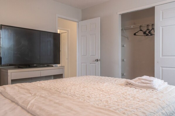Un dormitorio con cama, televisión sobre un mueble, armario con ropa colgada y una puerta que da a otra habitación.
