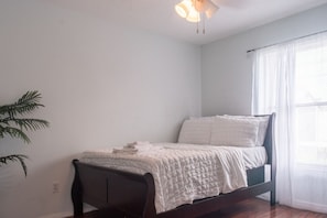 A single bed with a white bedspread and pillows, a dark wooden headboard, a ceiling light fixture, and a window with sheer curtains. - Port St. Lucie Poolside Oasis (Port St. Lucie)