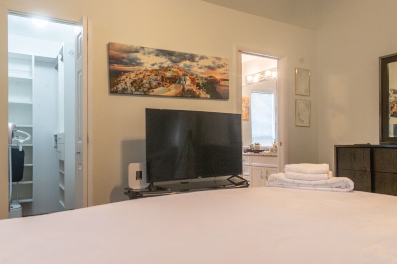 A bedroom with a bed, a flat-screen TV, a painting, a mirror, and a bathroom visible through an open door.