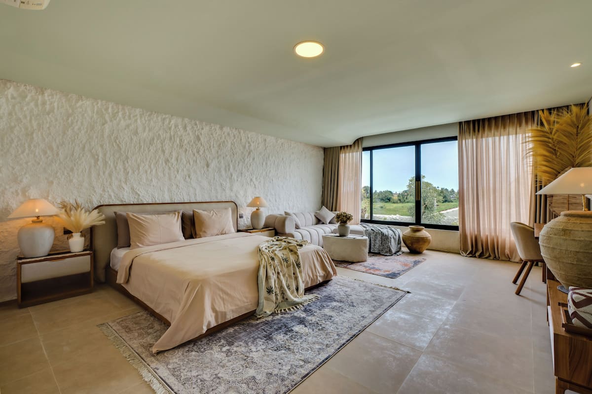 A bedroom with a large bed, bedside tables, a nightstand, a window with curtains, and a view of greenery outside.