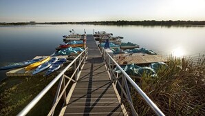 Une jetée en bois mène à un lac paisible, où des kayaks et des canoës sont amarrés sur la rive droite.