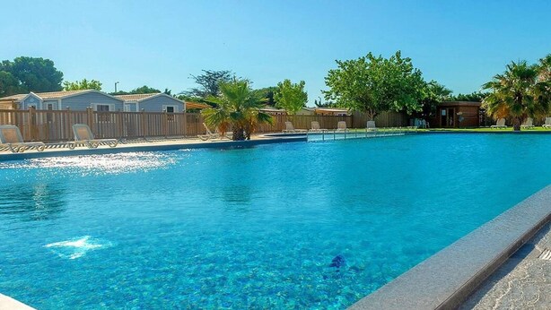 Una piscina cristallina con recinzione in legno, sedie a sdraio e una palma.