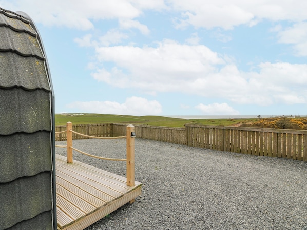 A wooden deck with a rope barrier, gravel ground, and a view of green hills and the sea. - Keith (Stranraer)