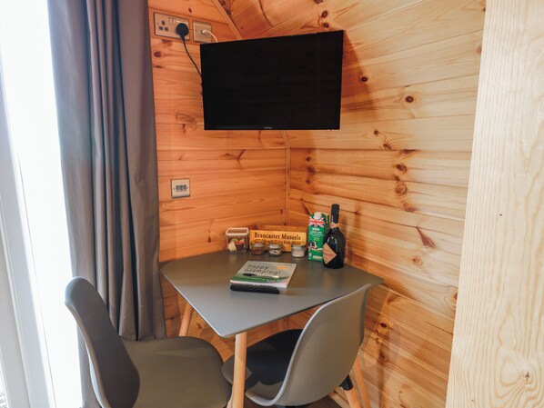 A small cabin with a table, two chairs, a television mounted on the wall, and a window with curtains.