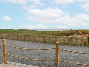 A wooden fence with rope barriers, a gravel path, and a view of green hills and the sea. - Keith (Stranraer)