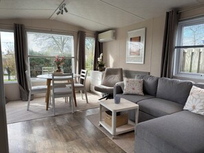 A modern living room with a grey sofa, white armchair, and dining table with chairs. There are large windows with curtains, a white air conditioning unit, and a framed picture on the wall. - Beautiful chalet with private parking space and quietly situated on the edge of our park. (Sint Maarten)