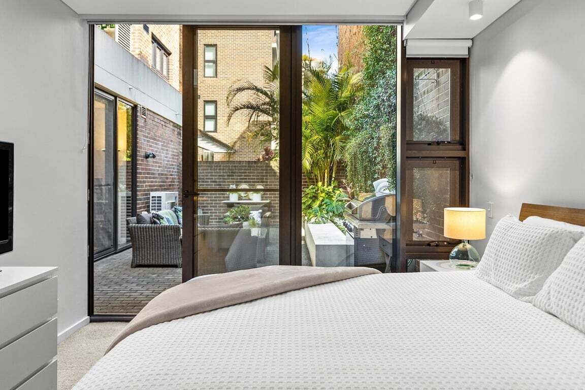 Une chambre à coucher comprenant un lit, une table de chevet, une lampe et donnant sur une cour intérieure avec une table et des chaises.
