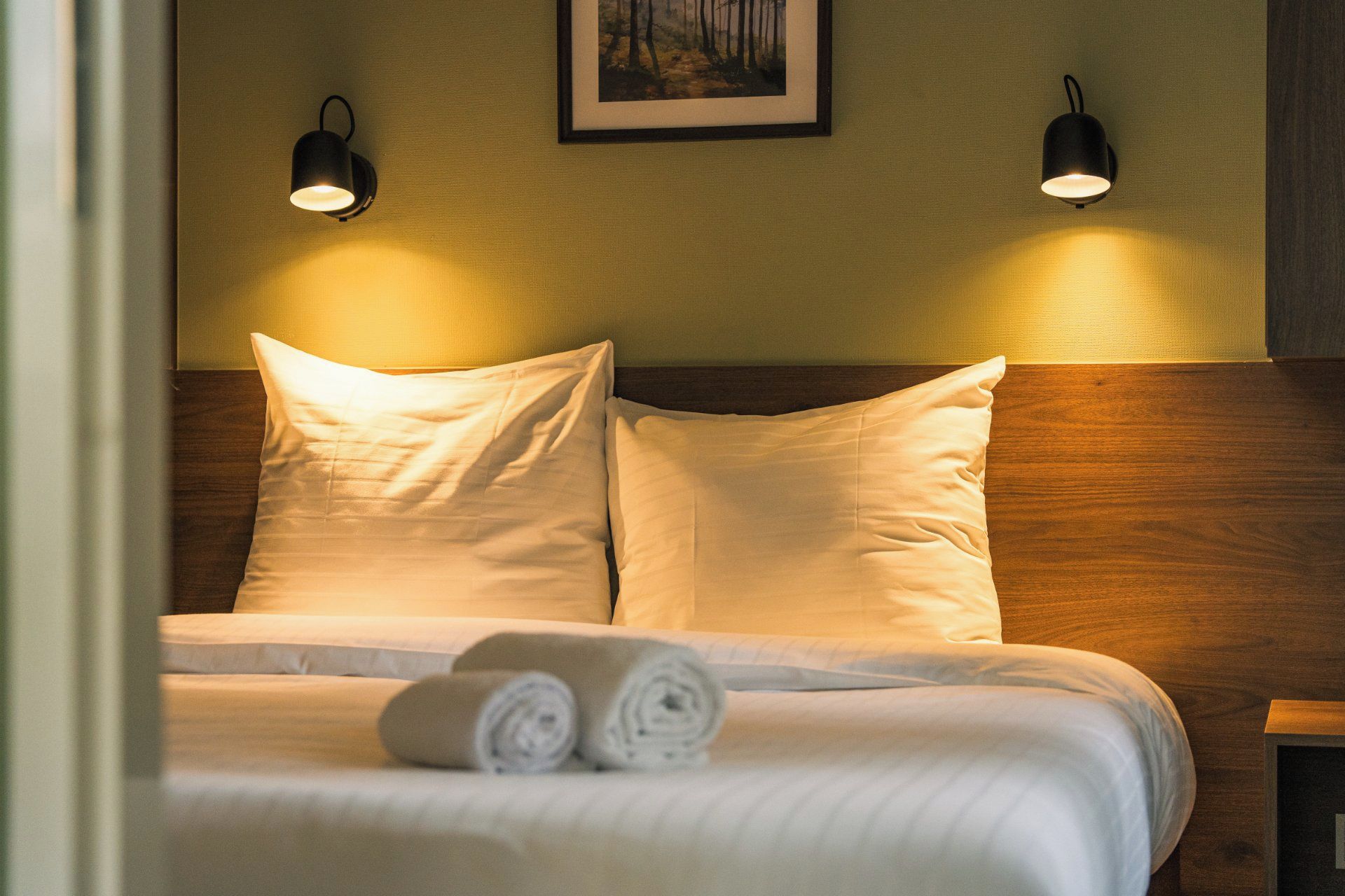 A neatly made bed with pillows and rolled towels, two wall-mounted lights, and a framed picture on the wall.