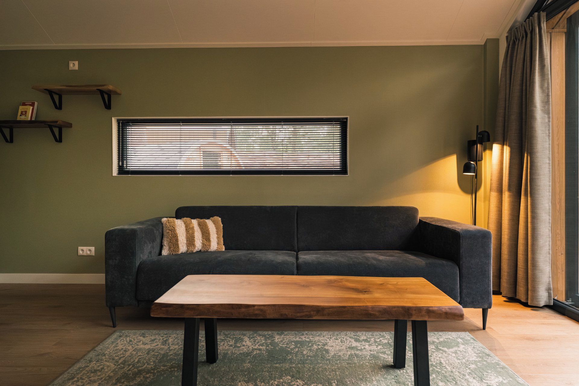 Un salon avec un canapé foncé, une table basse en bois, un tapis à motifs, une fenêtre avec des stores et une lampe.